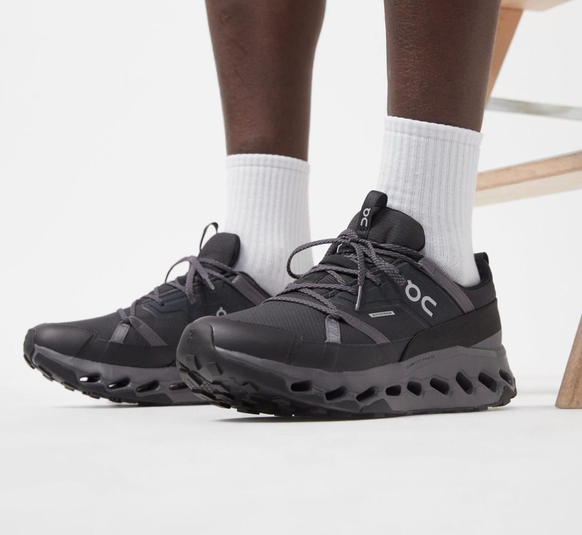 Close-up of a person wearing black On running shoes with gray laces and white crew socks, standing on a white floor.
