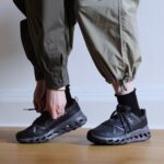 Person bends down to tie the laces of black athletic sneakers with a chunky, hollowed sole, wearing olive cargo pants.