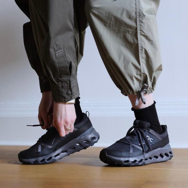 Person bends down to tie the laces of black athletic sneakers with a chunky, hollowed sole, wearing olive cargo pants.