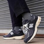 Close-up of grey and black New Balance sneakers with white laces, worn with dark jeans, on a city sidewalk.