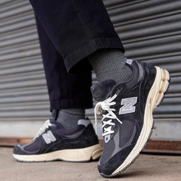 Close-up of grey and black New Balance sneakers with white laces, worn with dark jeans, on a city sidewalk.