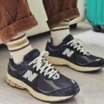 Person wearing black New Balance sneakers with cream soles, cream laces, and striped socks, standing on a light floor with rolled brown pants.