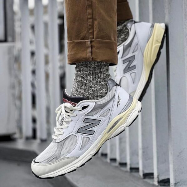 Person wearing white/gray New Balance sneakers with chunky sole, gray knit socks, and rolled brown pants leaning on a metal railing