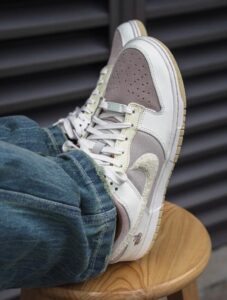 White and beige sneakers with perforated toe resting on a wooden stool, denim jeans visible at the cuff.