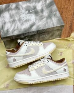Two white and gray sneakers with beige detailing and a rabbit motif on the heel, pictured beside a metallic shoebox.