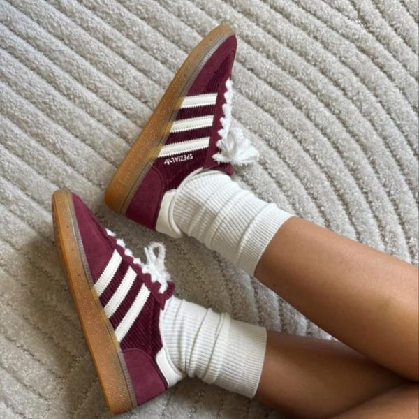 Pair of maroon sneakers with white Adidas stripes and gum soles, worn with white ribbed socks, resting on a light beige carpet.