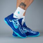 Blue high-top sneakers with turquoise soles featuring a bone-drawn design, worn with white crew socks and a tattooed leg showing above.