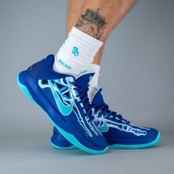 Blue high-top sneakers with turquoise soles featuring a bone-drawn design, worn with white crew socks and a tattooed leg showing above.