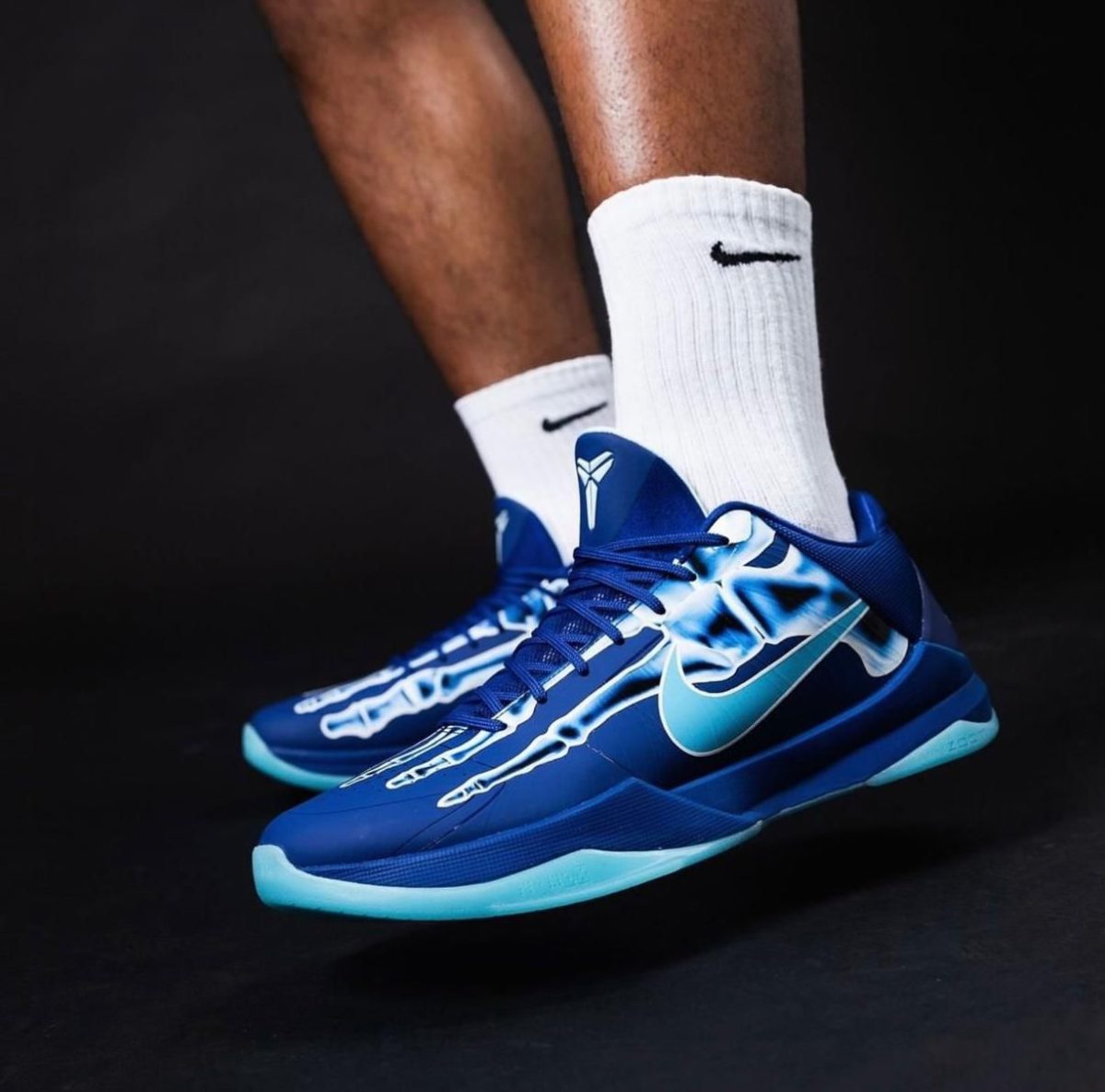 Blue athletic sneakers with turquoise accents and white socks featuring the Nike swoosh, photographed mid-step against a dark background.