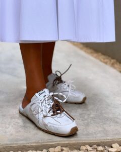 WhatsApp_Image_2024-12-30_at_13.02.40 | UAE SPORT STORE Close-up of white sneakers with brown laces worn with a white skirt, standing on a concrete surface.