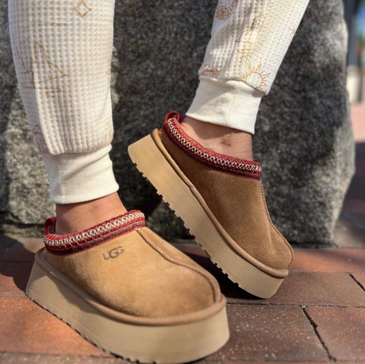 Tan suede UGG-style platform slippers with red woven trim, worn with cream knit pants on brick pavement.