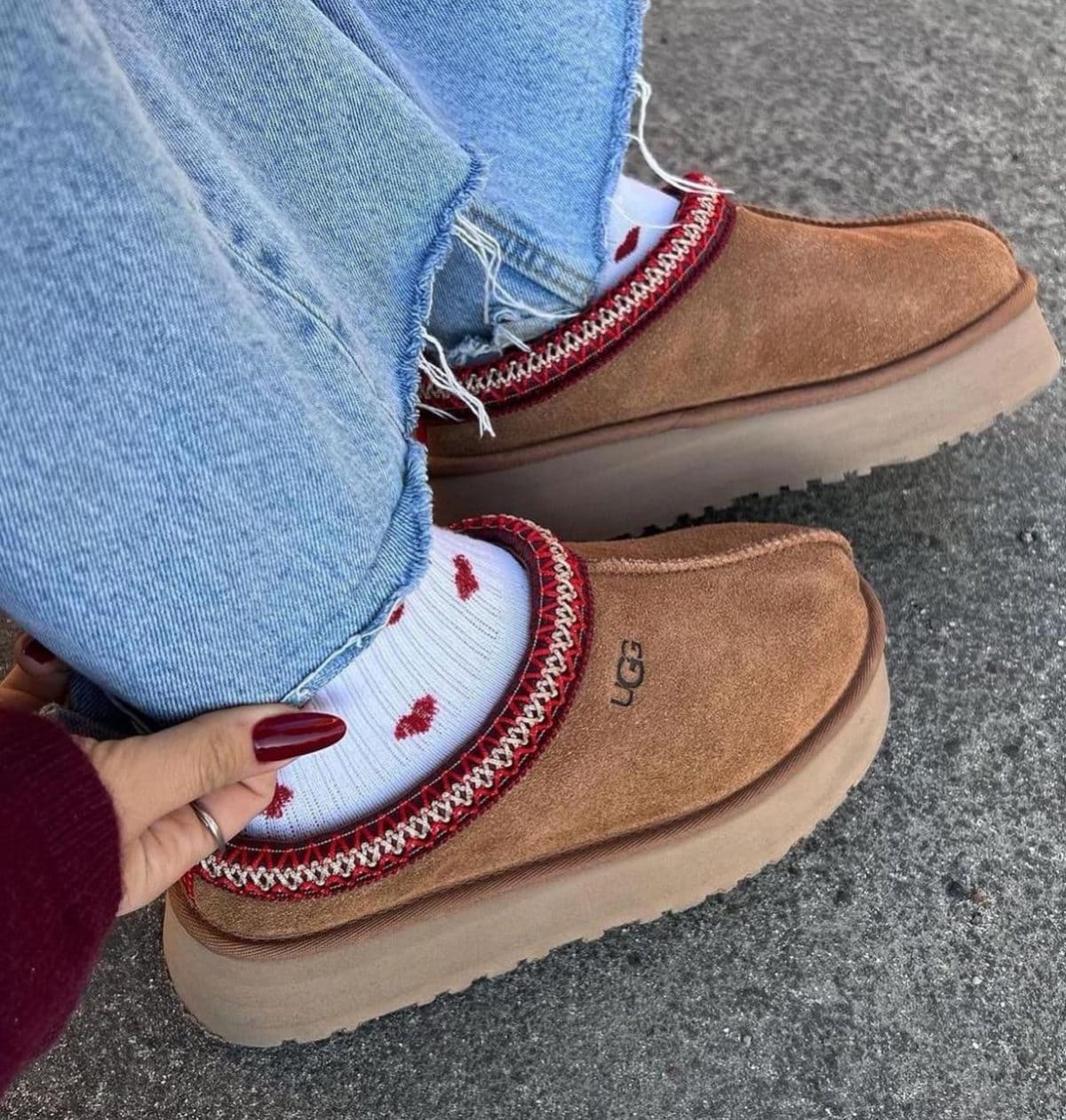 Person wearing tan suede moccasin-style shoes with a thick beige sole, paired with light-wash jeans and white socks with red lips, adjusting the fit.