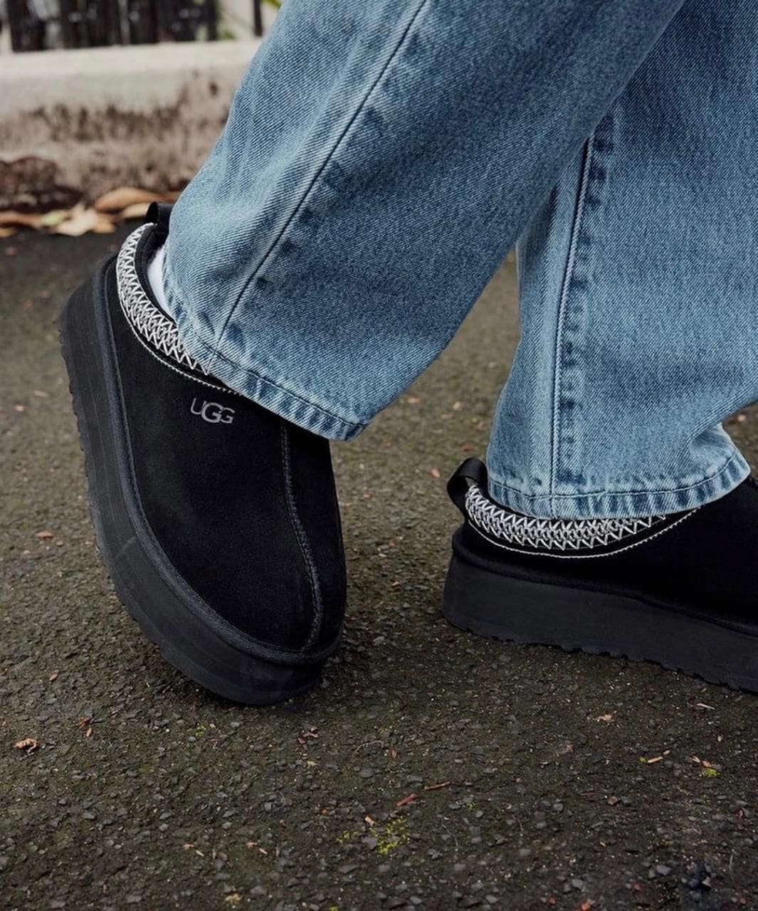 WhatsApp_Image_2025-01-10_at_12.27.46 | UAE SPORT STORE Close-up of black UGG boots with white decorative stitching, paired with light-wash jeans, stepping on a wet pavement outdoors.