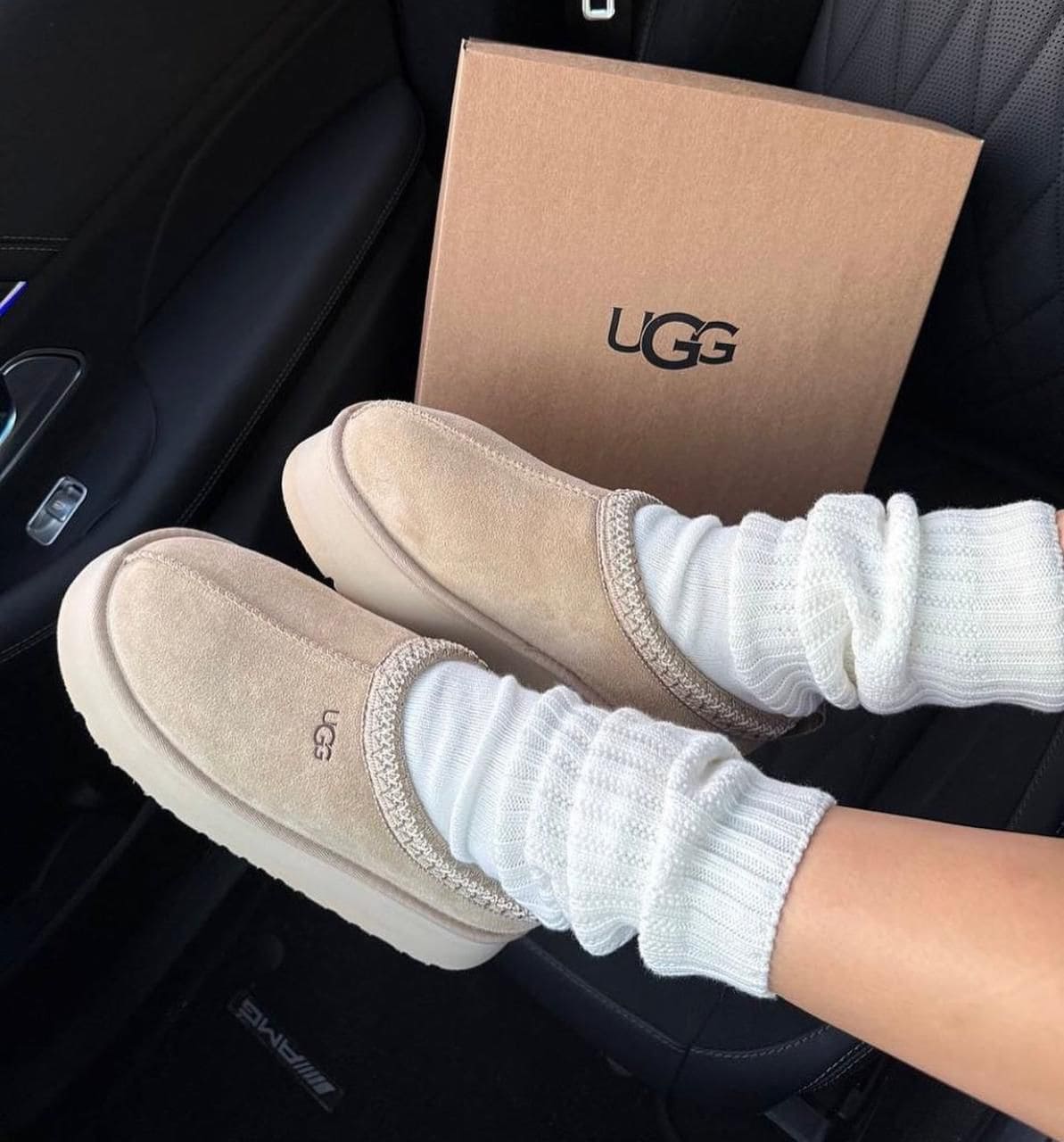 WhatsApp_Image_2025-01-10_at_15.30.28 | UAE SPORT STORE Beige UGG slip-on slippers on white socks, resting on a car seat with a UGG box in the background.