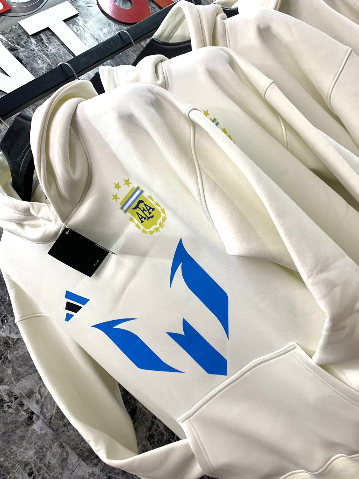 WhatsApp_Image_2025-01-10_at_18.38.56_1 | UAE SPORT STORE White hoodie on a hanger with a blue geometric logo and a small yellow crest with stars on the chest area