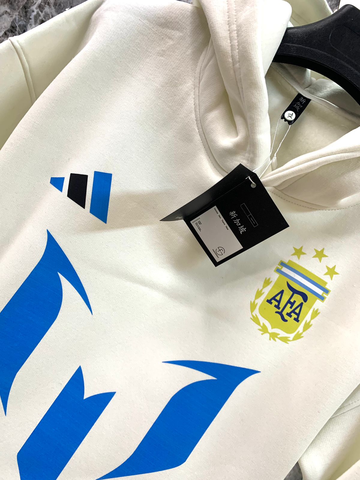 WhatsApp_Image_2025-01-10_at_18.38.57 | UAE SPORT STORE White hoodie on a hanger with blue geometric graphics and the Argentina Football Association (AFA) crest; black tags attached, size XL visible.