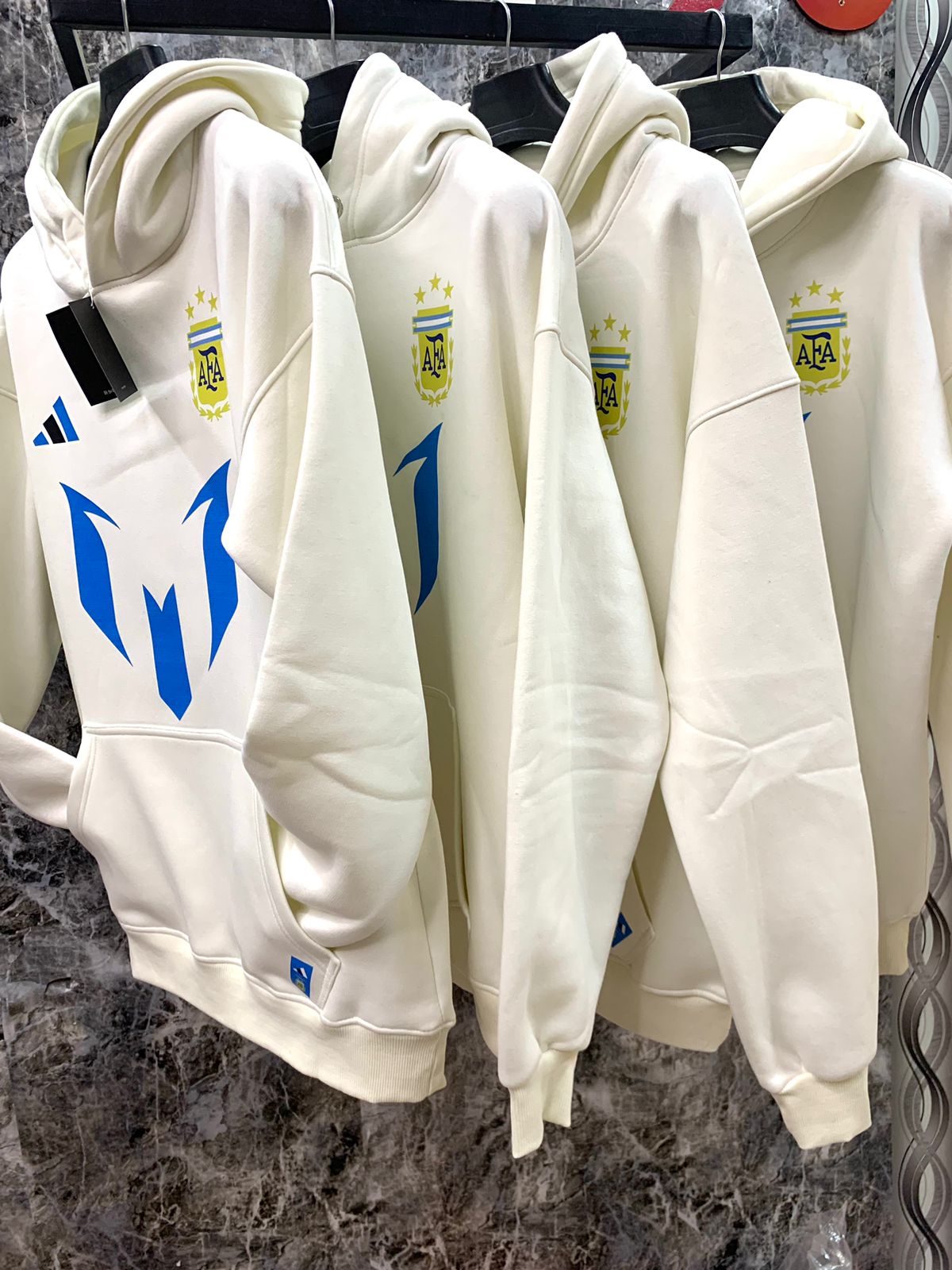 WhatsApp_Image_2025-01-10_at_18.38.57_2 | UAE SPORT STORE Rows of white hoodies on a clothing rack, each featuring a blue abstract emblem on the front and a small crest with stars on the chest surface.