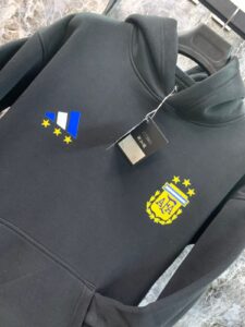 Black hoodie on a hanger with AFA crest and blue-yellow emblem visible; tag attached.
