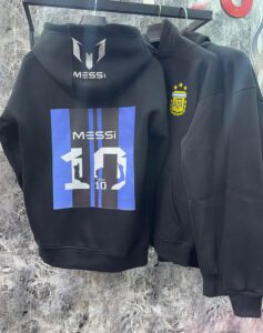 Two black hoodies on a hanger; the left hoodie has a blue vertical stripe graphic with 'MESSI' and the number 10 on the back, the right hoodie has a yellow crest with stars on the chest.