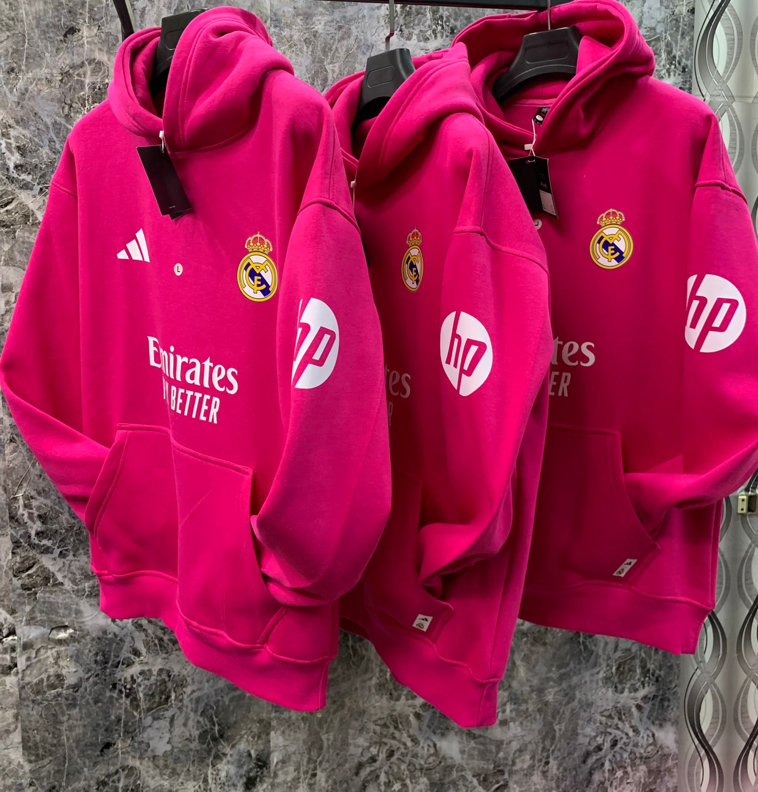WhatsApp_Image_2025-01-10_at_18.40.05 | UAE SPORT STORE Bright pink Real Madrid hoodies on hangers, showing club crest and sponsor logos Emirates and HP against a marble backdrop.