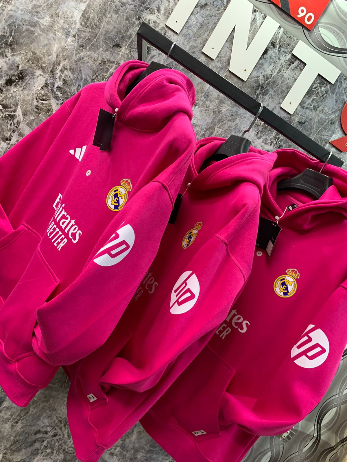 WhatsApp_Image_2025-01-10_at_18.40.06_1 | UAE SPORT STORE Bright pink Real Madrid hoodies on a display rack, showing team crest and sponsor logos Emirates and HP against a marble wall.
