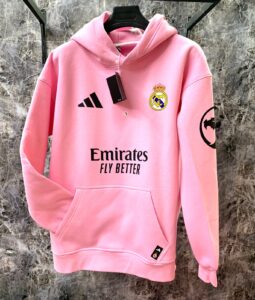 Pink Real Madrid hoodie on hanger with crest, Adidas logo, Emirates sponsor text, and a black tag visible on the chest area, against a marble background.