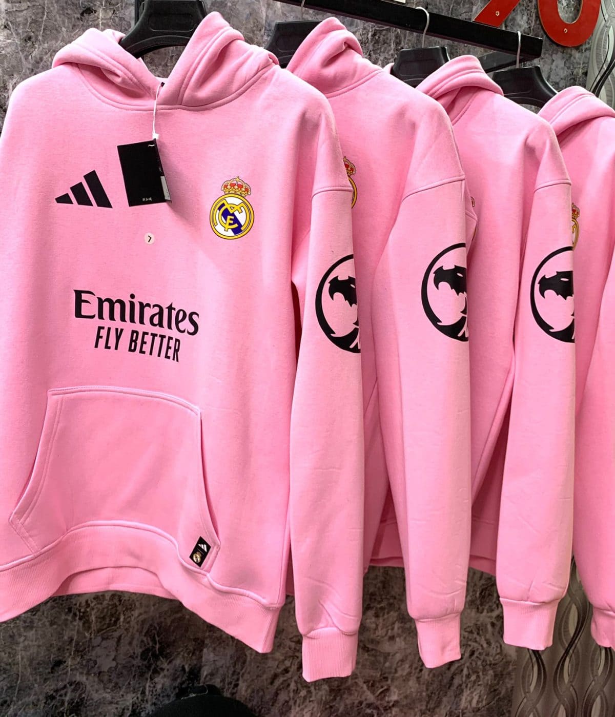 Pink Real Madrid hoodie on a rack featuring Emirates Fly Better slogan and team crest, with a black tag attached.