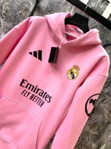 Pink Real Madrid hoodie on a hanger, featuring Adidas logo, Real Madrid crest, Emirates Fly Better sponsor text, and a Batman emblem on the sleeve.