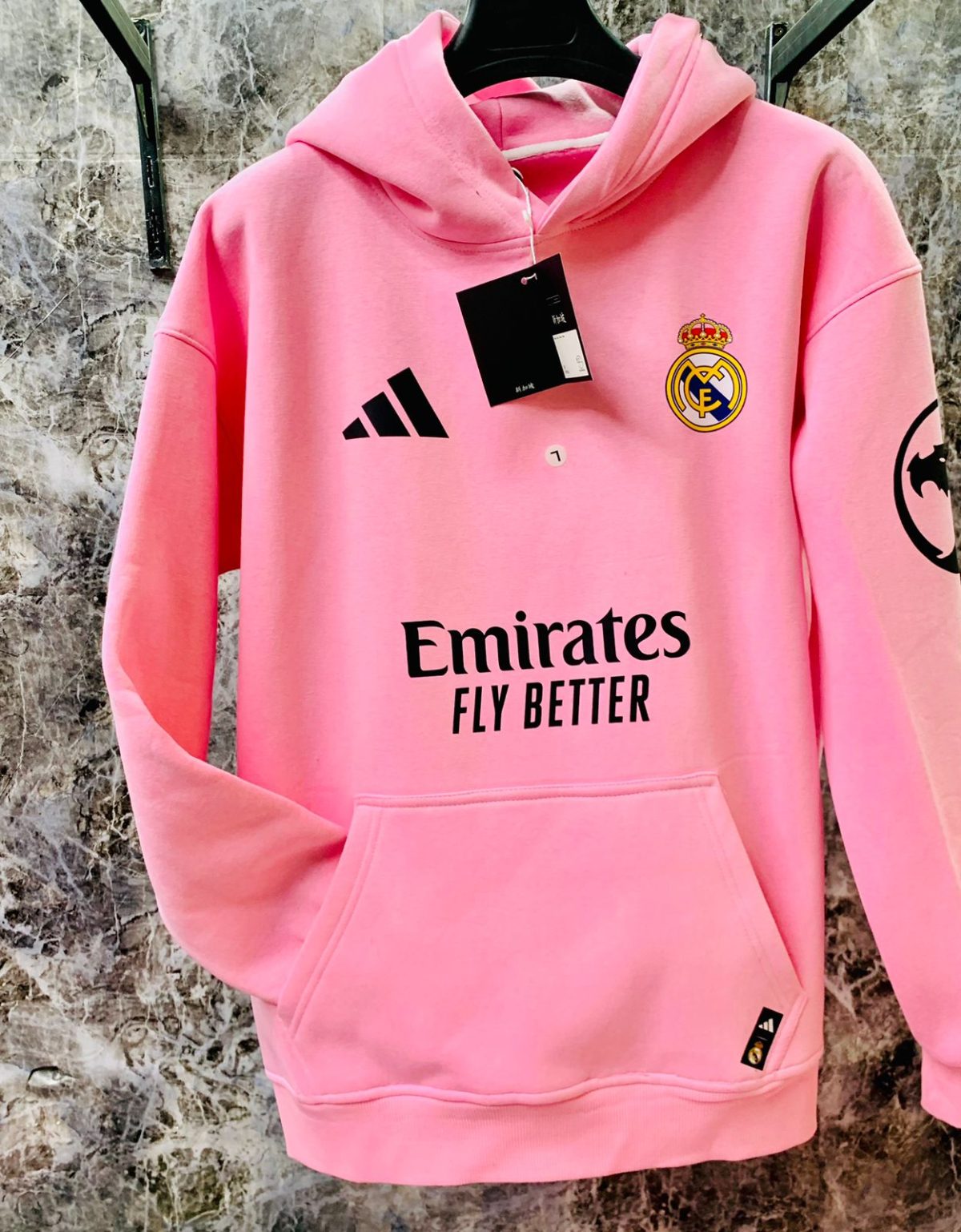 Pink Real Madrid hoodie hanging with club crest on the left chest and Emirates Fly Better slogan.