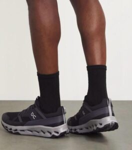 Person wearing black socks and dark gray On running shoes, standing with heels slightly elevated, showing the shoe sole detail.