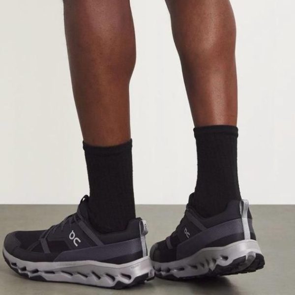 Person wearing black socks and dark gray On running shoes, standing with heels slightly elevated, showing the shoe sole detail.