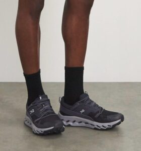 Person wearing black socks and dark gray athletic shoes standing on a light floor, showing the soles with a pronounced scalloped sole pattern (On logo on the sides).