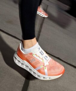 Person wearing coral running shoe with white sole on a sidewalk; black leggings and white sock visible inside the shoe.