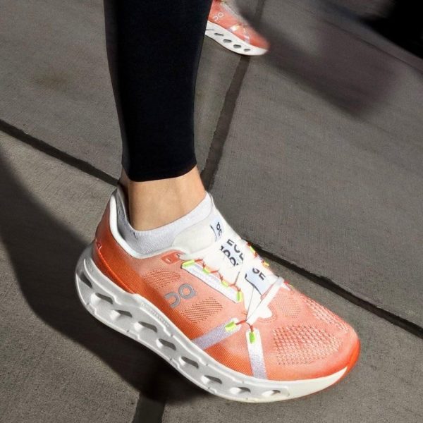 Person wearing coral running shoe with white sole on a sidewalk; black leggings and white sock visible inside the shoe.