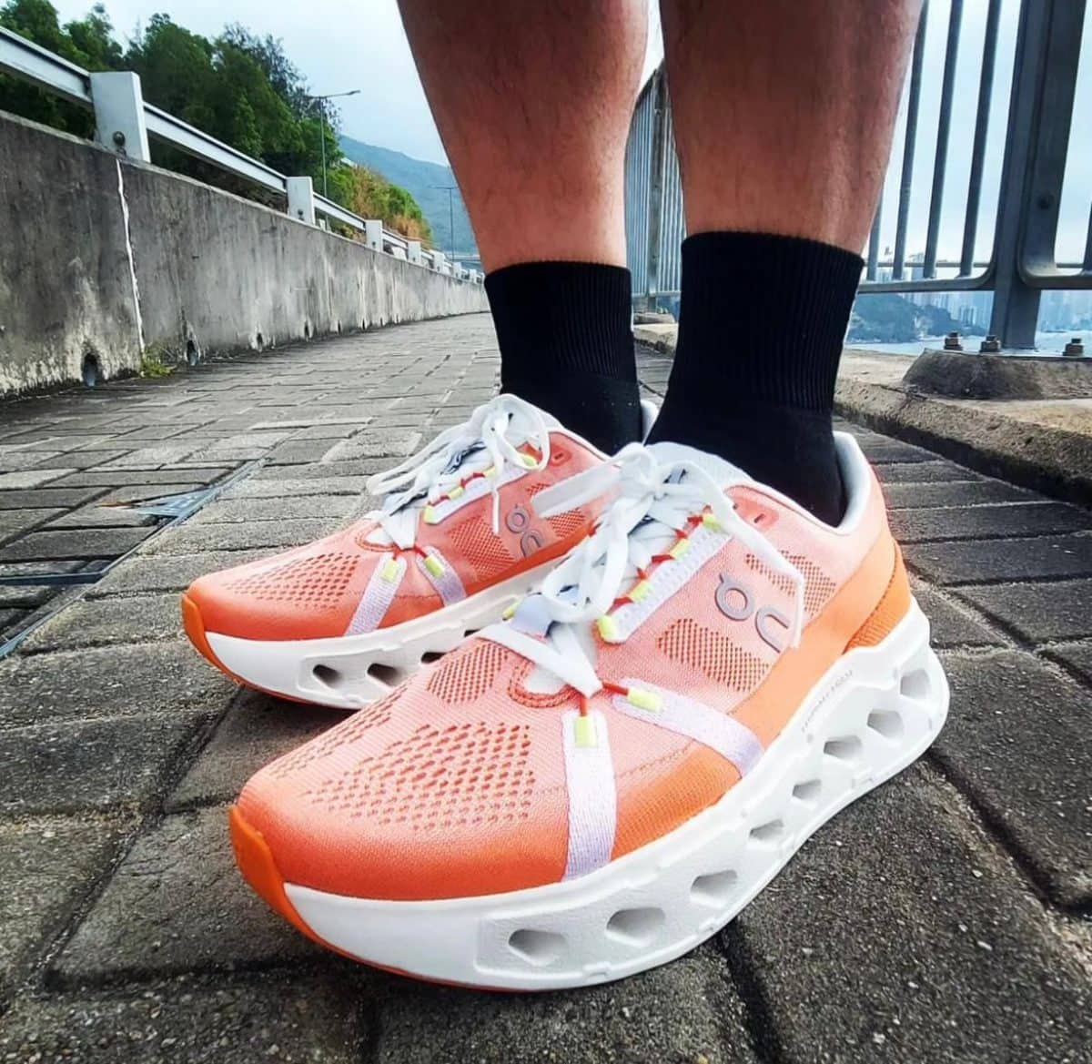 Peach-colored running shoes with white laces and a hollow white sole on a brick walkway beside a railing by the water.