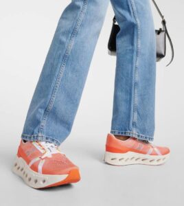 Person walking in bright orange sneakers with a white sculpted sole, paired with wide-leg jeans and a small black crossbody bag behind.