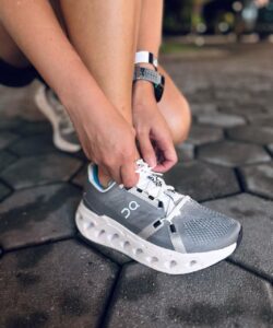 Person ties white laces on a gray On running sneaker while kneeling on a cobblestone pavement.