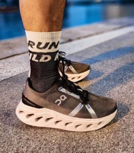 Foot wearing a dark gray On running shoe with a thick white sole, black sock, and a white sock showing RUN text, standing on a paved surface near a pool area.