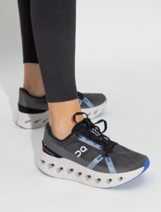 Person wearing gray and black running shoes with a white, hollowed-out sole and blue accents; On logo visible on the side.