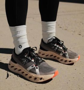 Gray athletic running shoes with breathable mesh, black accents, and orange-tipped soles worn with white crew socks on pavement.