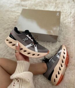 Gray On running shoe with peach sole held over a matching box on a white fluffy rug.