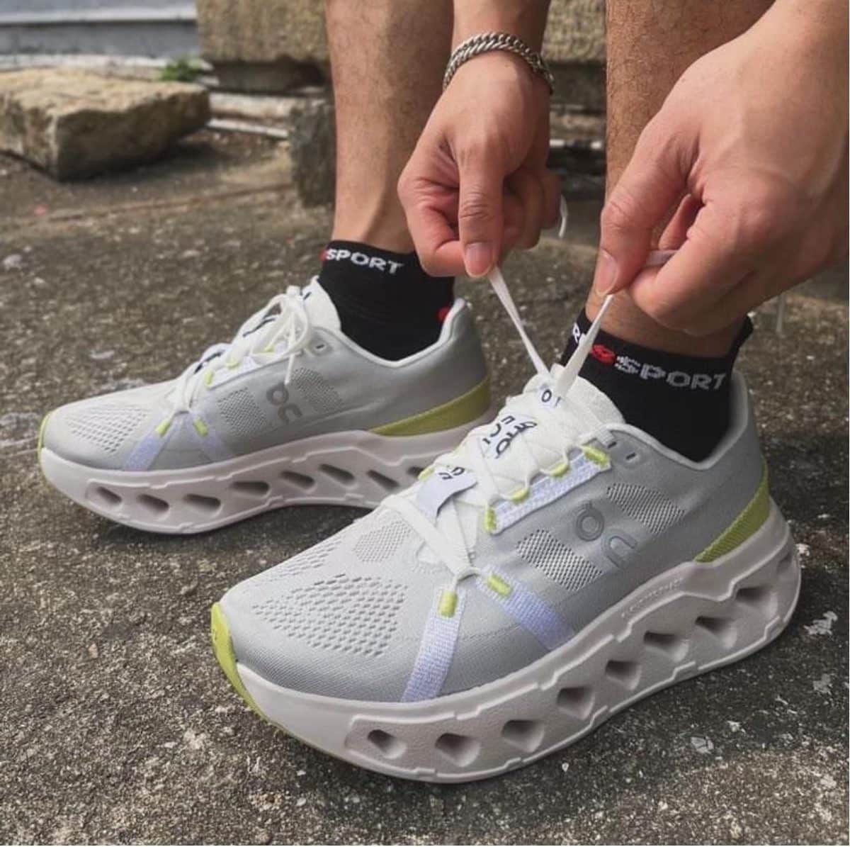 Person tying the laces of white running shoes with neon yellow accents on a concrete surface.
