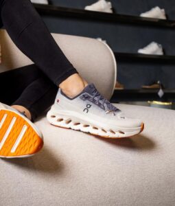 WhatsApp_Image_2025-01-22_at_13.03.22 | UAE SPORT STORE Close-up of a gray On running sneaker with an orange sole resting on a beige sofa, worn by a person seated indoors.
