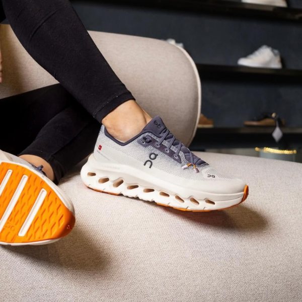 Close-up of a gray On running sneaker with an orange sole resting on a beige sofa, worn by a person seated indoors.