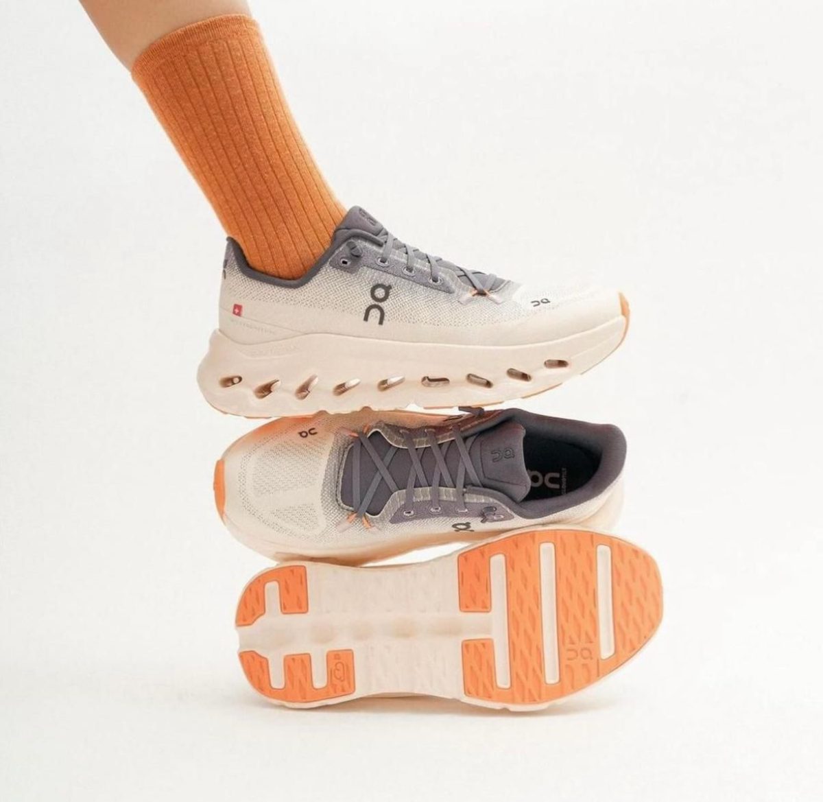 Pair of On running shoes in gray and orange, stacked mid-air with an orange sock visible above.