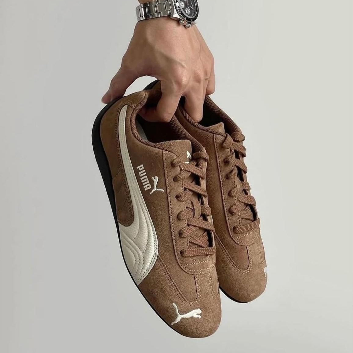 WhatsApp_Image_2025-01-28_at_10.04.04 Brown suede Puma sneakers with white logo held by a hand wearing a silver watch, against a neutral background.