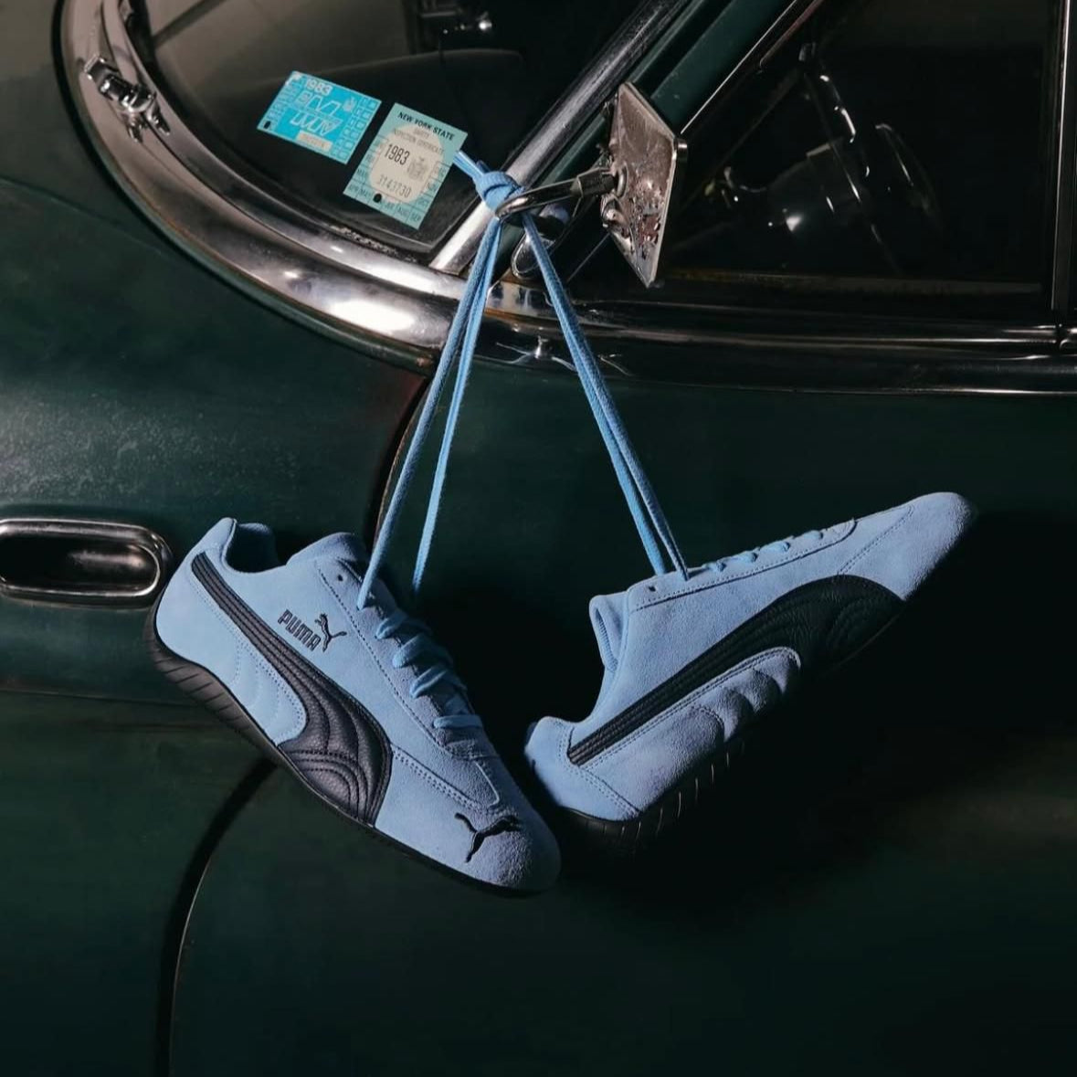 WhatsApp_Image_2025-01-28_at_15.59.30 Light blue Puma sneakers hanging by their laces from a vintage dark-green car door.