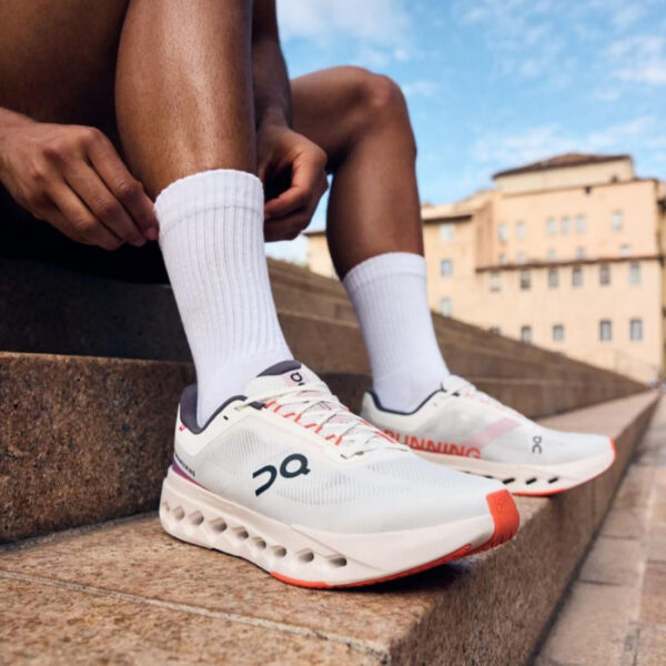 Close-up of white running shoes with orange soles worn by a person sitting on stone stairs, urban buildings in the background.