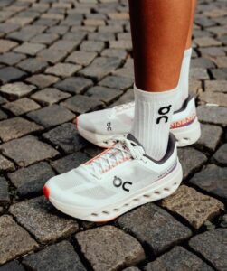 White running shoes with black 'bc' logo being worn over white socks, standing on cobblestone pavement.