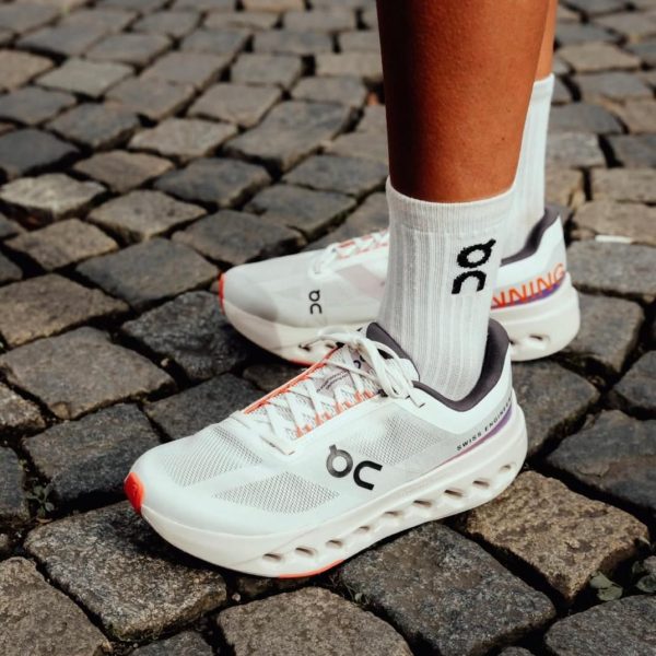 White running shoes with black 'bc' logo being worn over white socks, standing on cobblestone pavement.